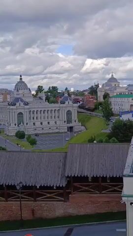 The media file captures a sweeping aerial view of Kazan, Russia. In the foreground, a weathered wooden structure with a tiled roof is visible, suggesting it might be part of an older defensive wall or historical building. Beyond this, the scene opens up to reveal a wide expanse of the city.

The main focus is on a grand, white, neoclassical building with a prominent dome, likely a public building or a palace, situated along a tree-lined avenue. Cars are seen on the road and in a parking lot adjacent to the building, indicating a normal day with people visiting or working there.

Further back, a lush green park with walking paths and scattered trees offers a recreational space. Beyond the park, a vast body of water, likely the Kazanka River or Kuybyshev Reservoir, stretches out, reflecting the sky. Across the water, a modern cityscape unfolds with numerous high-rise buildings forming a distinct skyline. The contrast between the historical architecture in the mid-ground and the contemporary skyscrapers in the distance highlights the city's blend of old and new.

The sky is dramatic, filled with a mix of heavy, grey clouds and patches of bright blue, suggesting a dynamic and possibly changeable weather. The lighting indicates it is daytime, likely late morning or early afternoon. No people are clearly identifiable as distinct subjects, though they can be seen as small figures in the park and on the avenue. There is no visible text in the media file. The overall impression is one of a well-developed city with significant historical and modern elements, set against a natural landscape.