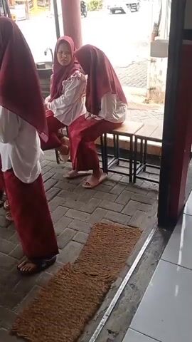 The video shows a group of four girls, all wearing maroon hijabs and maroon skirts with white tops, standing in an outdoor area with a brick-like floor. Two of the girls are sitting on a low bench to the right, while the other two are standing. The girl on the far left is partially visible from the side, while the girl next to her is facing the camera. The girl on the far right is facing away from the camera, looking to her right. The girl in the middle, standing, has her hands near her face, and appears to be looking down or crying. The overall scene suggests a break or waiting period, possibly at a school or an event, given their uniform attire. In the background, a street with a truck and a motorcycle can be seen. There are also food stalls visible on the left side of the frame.