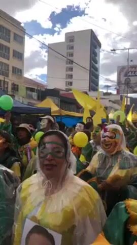 The video shows a street celebration with a large crowd of people dressed in green and yellow costumes. Many are wearing masks or face paint. They are carrying yellow flags and balloons. The crowd appears to be moving down the street. The buildings on either side of the street are visible in the background, along with a large billboard advertising a political candidate. The weather appears to be overcast.