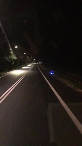 A first-person perspective video shows a road at night with streetlights illuminating the path ahead. A blue object with lights is visible on the right side of the road. The camera is moving forward, suggesting travel by vehicle or bicycle.