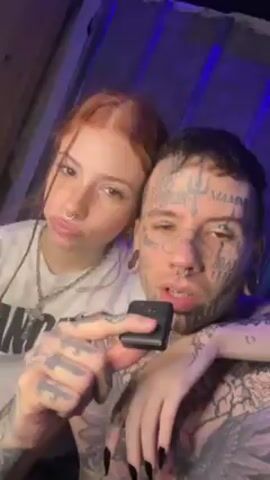 Here's a description of the video:
A close-up shot shows a young couple. The woman has light skin, red hair pulled back in braids, a nose ring, and is wearing a white t-shirt. The man has tattoos covering his face, neck and arms. He has multiple facial piercings and stretched earlobes. He is holding a small black device close to his mouth. The woman has her arm around the man's shoulders. They are speaking to the camera in different languages. The background is a dark shade of blue and appears to be inside a room.