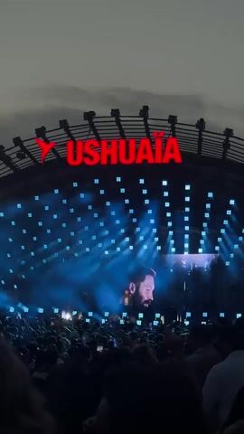 A live performance is taking place at Ushuaia, a venue indicated by a large red sign. A large screen displays a close-up of a man with a beard, possibly the performer. The stage is lit with numerous blue lights, creating beams that project into the darkness. A large crowd of people is visible in the foreground, looking towards the stage. The sky is dark and appears cloudy. The video shows a dynamic light show and the enthusiastic crowd.