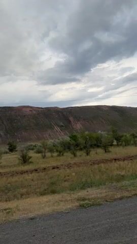 The video shows a view of a large hill or mountain range, possibly a mine or quarry, with reddish-brown soil and patches of green vegetation. The sky is overcast with dramatic clouds. In the foreground, there is a dirt road or path, and some sparse vegetation and trees. A utility pole is visible on the right side of the frame. The camera is panning slowly to the right. The overall scene suggests an outdoor, possibly rural or industrial, environment. The audio consists of music.