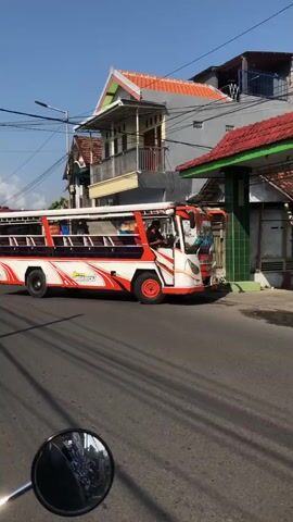 A low-angle shot shows a white bus with orange and red stripes driving down a street. The bus is open on the sides with benches for passengers. The bus has "SUPER PREMIUM" written on the side in white lettering with a graphic of people sitting on benches. A person in a red shirt is visible inside the bus. The bus is driving on a paved road, and there are houses and power lines in the background. The sky is blue and clear. The video appears to have been filmed from a moving vehicle, as a motorcycle mirror is visible in the bottom left corner.