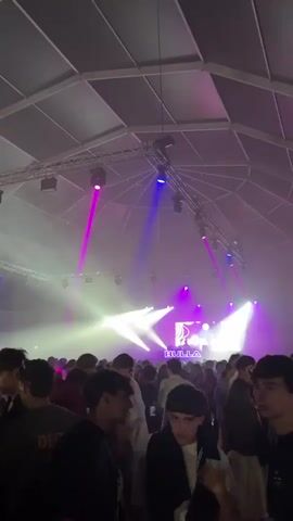The video shows a crowded indoor event with a large number of young people. The venue appears to be a tent or a large hall with a white, domed ceiling supported by a metal structure. Lighting is a prominent feature, with beams of pink and blue light illuminating the space and creating a smoky atmosphere, likely due to fog or haze machines. In the background, a stage or DJ booth is visible with bright lights and a logo that reads "BULLA". The crowd is dense, with people standing and milling around. Most individuals appear to be in their late teens or early twenties. Some are looking towards the stage, while others are interacting with each other. The overall impression is of a lively party or concert.