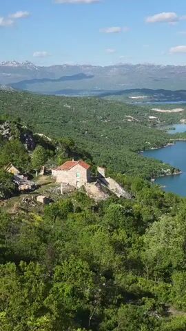 The video shows a scenic view of a landscape with a lake, green vegetation, and mountains in the background. A small house with a red-tiled roof is visible on a slope covered in trees. The lake is expansive, with several small, tree-covered islands dotting its surface. The mountains in the distance have visible snow on their peaks. The sky is bright blue with scattered white clouds.