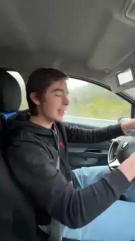 A young man is driving a car on a gray day. He is wearing a black hoodie with a small logo and light blue jeans. He looks at the camera and gestures while he talks. The road outside the window is gray and wet. He is wearing his seatbelt.