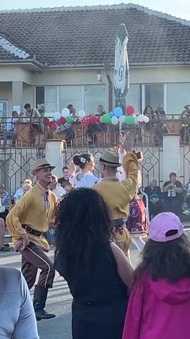 A group of people are gathered outdoors for what appears to be a cultural event or celebration. In the foreground, two dancers in traditional attire are performing. The male dancers wear hats, yellow shirts with intricate trim, and dark pants tucked into boots. One dancer is facing forward, his left arm raised and his right hand extended. The other dancer is facing away, his left arm raised and his right hand touching the first dancer's hat. Behind them, two female dancers are also present, one in a white blouse and skirt with a floral pattern, the other in a white blouse and a dark skirt with a floral pattern. They are holding hands and appear to be part of the dance. Several spectators are visible in the background, seated on chairs and standing on a balcony. The building behind the dancers has a balcony decorated with balloons in various colors, including red, white, green, and blue. The environment suggests a daytime event, with sunlight casting shadows and illuminating the scene.