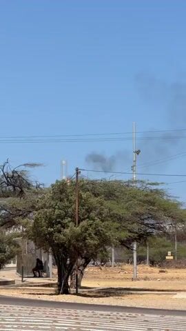 The video shows a landscape under a bright, clear blue sky. High winds are audible throughout the recording. In the foreground, there is a roadside bus stop where a person is sitting. There is a tree between the bus stop and the road. In the background, an industrial facility is visible. A tower is expelling a large plume of orange fire and black smoke. The road in the foreground curves towards the facility. Power lines and poles are visible overhead. The ground is dry and sandy with sparse vegetation.