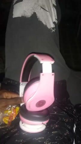 The video shows a close-up of a pair of pink and white headphones being held by a person's hand. The headphones are being manipulated, with one ear cup being turned inward. The person is wearing a black t-shirt with a white graphic design and dark-patterned fabric is visible in the background. The lighting is dim, suggesting an indoor setting at night.