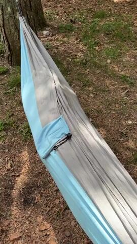 A young woman with long blonde hair lies in a hammock in a forest setting, looking up at the camera. She is wearing a white t-shirt and denim shorts. The hammock is light blue and gray, with a small pouch on the side. The ground is covered in brown leaves and green grass. A tree trunk is visible in the background. The video shows her briefly appearing and disappearing from the hammock.