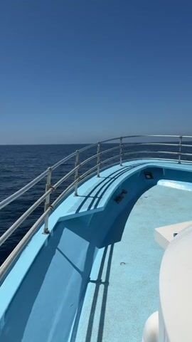 This is a first-person perspective video of a boat moving through calm, blue ocean waters under a clear, bright blue sky. The foreground shows the blue deck of the boat with a metal railing along the edge. The railing is casting shadows on the deck. The vast expanse of the ocean stretches out to the horizon where it meets the sky. The boat appears to be moving forward at a moderate pace, indicated by the slight movement of the water and the perspective. The lighting suggests it is daytime, likely midday due to the brightness. There are no people visible in the frame. The scene is peaceful and evokes a sense of travel and open water.