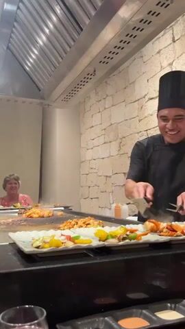 A chef in a black chef's hat and black uniform is cooking on a flat top grill in front of a stone wall. He is smiling and using two spatulas to flip food on the grill. In the background, a woman in a pink shirt is seated at the grill. Plates of food, including shrimp and vegetables, are arranged on the counter in front of the grill. There are also small condiment dishes in the foreground. The environment appears to be a restaurant with a stainless steel ventilation hood above the grill.