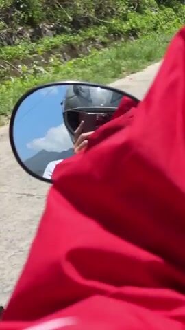 The video appears to be shot from the perspective of someone riding a motorcycle on a road. A rearview mirror reflection shows a person wearing a helmet and red jacket, holding a smartphone. The motorcycle is moving along a road, with lush green foliage lining the sides. The view expands to reveal a scenic mountain range in the background, under a clear blue sky. Utility poles with power lines can be seen along the road.