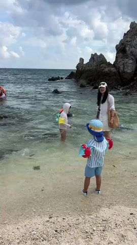 A group of people are enjoying a day at the beach with a rocky coastline and cloudy sky. In the foreground, a child wearing a blue and white striped shirt, blue shorts, a blue and yellow hat, and arm floaties stands on a sandy beach facing the ocean. The child holds a blue and yellow water squirter. In the middle ground, a woman in a white shirt and skirt, wearing a white hat, stands in the shallow water. She is looking towards the child. Another child in a light-colored outfit and a white hooded swimsuit is also in the shallow water. Further out in the ocean, a person in an orange life jacket is partially visible. The water is clear and shallow near the shore, with rocks visible both in the water and on the coastline.