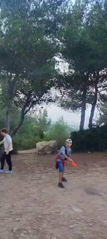 The video captures a young boy playing frisbee outdoors. He is in the foreground, wearing a blue t-shirt, shorts, and a white baseball cap. A red frisbee is visible in the air above and slightly in front of him. The background shows a natural environment with trees and a dirt path. The lighting suggests it might be late afternoon or early evening. The boy appears to be mid-throw or preparing to catch the frisbee. The scene is casual and appears to be a leisure activity in a park or wooded area.