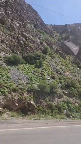 The video shows a mountainous landscape on a sunny day. The camera is moving along a road that runs parallel to a mountain. The mountain is covered in green vegetation and rocky areas. An electric power tower is visible on the slope of the mountain. Power lines are seen running alongside the road. The sky is clear and blue with a few wispy clouds.