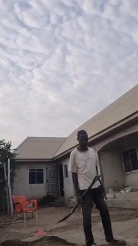 The video shows a man standing in a dusty yard, holding a digging tool with a long handle and a flat, curved blade. He is wearing a light-colored t-shirt, dark pants, and casual sandals. He uses the tool to dig into a pile of sand and mix it with water. In the background is a single-story house with a light-colored roof and several windows. An orange plastic chair sits near the house. Other objects around the yard include buckets and building materials. The sky is overcast.