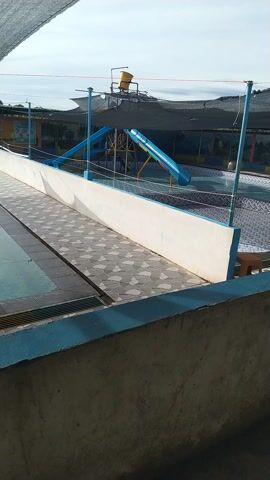 The media file shows a view of a swimming pool area, possibly at a water park, with a slide visible. The image is oriented vertically. A blue slide is prominent, positioned above the pool. There are metal structures supporting the slide and other equipment, painted blue and yellow. A yellow bucket-like fixture is attached to one of these supports. The pool area itself has tiled edges and appears to be empty of people. The water in the pool is a light blue color.

In the background, a dark mesh-like canopy stretches across a portion of the area, providing shade. Beyond the canopy, there are colorful images or murals painted on a wall. The sky is visible and appears bright, suggesting it is daytime. The overall lighting indicates fair weather. The tiling on the pool deck features a geometric pattern of light and dark grey diamond shapes. The edge of the pool is painted a bright blue. To the right of the pool area, a textured, grey concrete structure or wall is visible, casting a shadow.

No people are visible in the frame, and no specific activity is clearly taking place beyond the static presence of the water park amenities. The scene appears to be set in an outdoor location, likely a recreational facility. No legible text is clearly discernible in the media file. The location context provided is Meureudu, Indonesia.