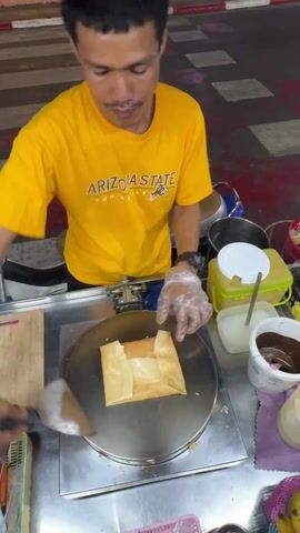 A man in a yellow Arizona State T-shirt is making a crepe at a street food stand. He uses a spatula to fold the crepe on a circular griddle. He then retrieves a square piece of paper from behind him. The stand is equipped with various food containers, including one filled with chocolate spread. The stand is set up on the street, with a scooter parked nearby and a crosswalk visible in the background.