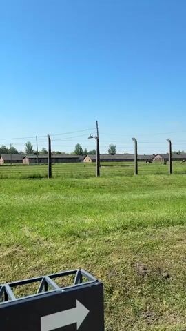 The video depicts the exterior of Auschwitz II-Birkenau concentration camp on a bright, sunny day.  The camera pans across a grassy field, showing the barbed-wire fences, electric posts, and lamps that defined the perimeter of the camp. In the background, rows of barracks are visible. A weathered stone pathway leads into the distance. A guard tower stands watch in the distance to the left. A short stretch of train tracks runs alongside the fence. An arrow sign is visible in the lower left corner. The scene is still and quiet.