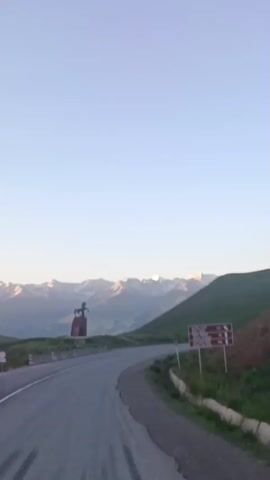 The video shows a curved asphalt road in a mountainous landscape. On the left side of the road, there is a monument of a horse on a pedestal. The monument is dark in color and is situated on a grassy area next to a concrete barrier. In the background, snow-capped mountains are visible under a clear blue sky. The lighting suggests it is either early morning or late evening. A red and white sign is visible on the right side of the road. The perspective suggests the video is being recorded from a moving vehicle.