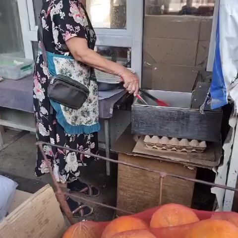 A woman wearing a floral dress and an apron is standing in front of a makeshift food stall. She is using tongs to remove a piece of fried fish from a metal container filled with oil. The fish appears to be golden brown and is being placed onto a tray. To the left of the woman, there is a table with some food items and containers. To the right, there is a wooden box with an egg carton on top, and a blue and white striped awning. In the foreground, there are some fruits, possibly oranges, in a basket. The scene suggests a street food vendor preparing to serve food.