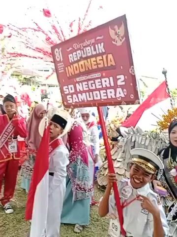 The video shows an outdoor gathering, likely a parade or festival, dominated by red and white decorations. In the foreground, several children are visible, dressed in uniforms and holding a large sign that reads "DIRGAHAYU REPUBLIK INDONESIA SD NEGERI 2 SAMUDERA". One child is carrying an Indonesian flag. The children are part of a larger procession, with many other people in the background, some wearing traditional attire and hats. The environment appears to be a grassy field with trees and buildings in the distance. The overall atmosphere seems celebratory, with a focus on Indonesian national pride. The audio includes a voice speaking in Indonesian, announcing "Finish-nya tetap di lapangan ini ya. Finish." which translates to "The finish is still in this field. Finish."