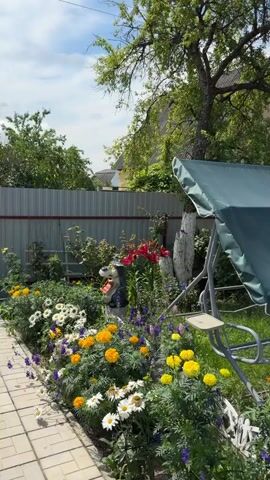 The video shows a garden with a swing set, a children's pool, and a trampoline. There are also several buildings, including a house with a red roof and a smaller wooden structure. The garden is filled with flowers and greenery. The weather is sunny with a few clouds in the sky.
