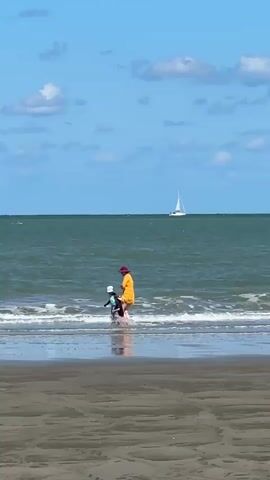 A person wearing a yellow shirt and a red hat walks in the shallow water at the edge of a sandy beach. A child in a blue swimsuit and a white hat stands to the left of the person, also in the shallow water. The ocean is a deep green, and small waves are breaking on the shore. In the distance, several sailboats with white sails are visible on the horizon. The sky is a bright blue with scattered white clouds. The camera is positioned at a low angle, looking towards the ocean from the beach.