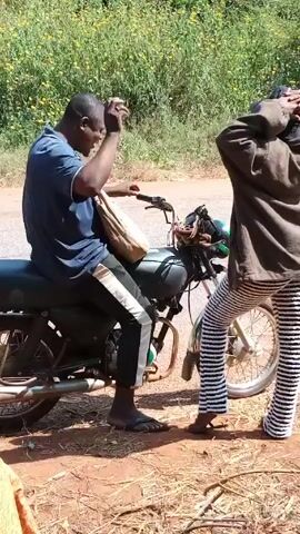 A man sitting on a motorcycle is looking into a brown bag. The motorcycle is on a dirt shoulder next to a paved road. A second person stands near the motorcycle, wearing striped pants and a brown jacket. There are weeds and flowering plants in the background.
