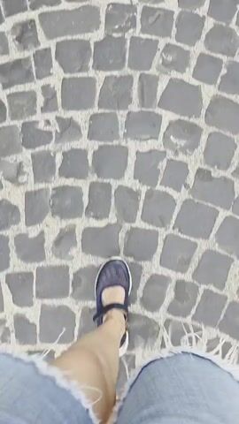 The video begins with a close-up of cobblestones as a person's feet, wearing sandals and denim shorts, walk forward. The perspective then shifts to a sped-up, blurry view of a cobblestone street with buildings on either side, suggesting movement through a town. The camera then stabilizes on a view looking up a narrow cobblestone street lined with buildings. In the distance, a large stone castle or fortress with a cylindrical tower is visible atop a hill, with green foliage and stone structures leading up to it. Several people are walking down the street, and some appear to be children playing. The buildings on either side of the street have multiple stories, windows, and some storefronts.