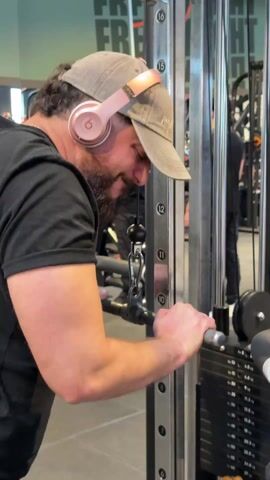 A man wearing a black t-shirt, gray baseball cap, and pink Beats headphones is exercising at a gym. He uses a machine with weights and a pull-down bar. The machine is marked with the brand name "MATRIX" and has numbered indicators from 8 to 16.  The man pulls the bar down. Other people are exercising in the background, and there is a sign with the word "FREE" visible. The setting is a well-lit gym.