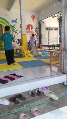 A boy in a teal shirt holds two balloons, one pink and one white, as other children play in a brightly decorated indoor playground. The playground features a yellow slide, a soft play structure with green and yellow steps, and a wall mural with cartoon animals and hot air balloons. A girl in purple is going down the slide, and another child is seated at the bottom. In the foreground, there is a mat on the floor, and several pairs of sandals are visible. The overall impression is of a fun, active environment for children.