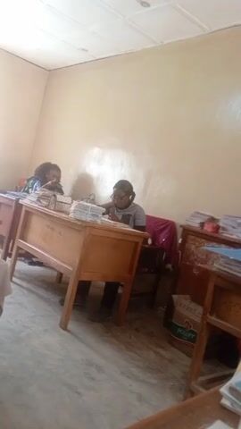 Two women are seated at desks in a modestly furnished room. The room has off-white walls, a tiled ceiling, and a cement floor. Each woman is surrounded by stacks of papers and appears to be working, possibly grading or reviewing documents. One woman wears glasses and has her hair pulled back. The other has curly hair. The desks are wooden and have writing on the front. A box labeled "Power Oil" is visible under one of the desks. The room seems to be an office or classroom setting.