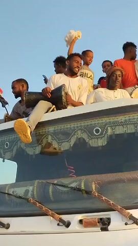 Here is a concise description of the video:

A group of people are joyfully celebrating atop a white truck. Several men sit on the truck's roof, one playing a drum energetically. Another man in a white robe leaps with enthusiasm. Children are interspersed amongst the men, waving and smiling. The sky above is a clear blue. The truck's windshield is visible, adorned with decorative tassels and rods.