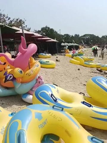 The video shows a beach with several inflatable tubes on the sand. In the foreground, there are large yellow and blue inflatable tubes. In the background, there are more inflatable tubes, some under canopies, and people walking on the beach and in the water. The sky is clear and the water is calm.