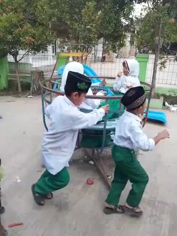 Several children, dressed in white shirts and green pants, are on and around a merry-go-round in an outdoor playground. Two girls in white hijabs are sitting on the merry-go-round, while three boys in black peci hats are pushing it to make it spin. Other playground equipment and trees are visible in the background. The playground appears to be located near a building with a green accent.