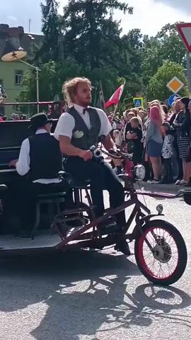 A man with long blond hair is seated on a bicycle, playing a horn. He is wearing a white shirt, a vest, and dark pants. Another man in a similar outfit, wearing a hat, is playing a piano. The piano is attached to the bicycle, and both men are part of a procession with a crowd of people watching. The background shows buildings and trees, suggesting an outdoor event.