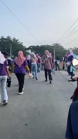The video shows a group of women walking along a paved road. Many of the women are wearing hijabs and purple shirts, suggesting they might be part of an organized group or event. One woman is riding a red scooter, facing away from the camera, and she is wearing a black headscarf and blue jeans. The environment appears to be outdoors, with trees and power lines visible in the background. The sky is a light blue, indicating it is daytime. The women are walking in the same direction, and the overall atmosphere seems to be one of movement and activity. There are no clear interactions between the individuals, beyond them sharing the same space.