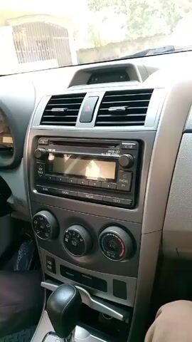 The camera shows the interior of a car, specifically the dashboard and front seats. The dashboard is a silver-gray color and features a radio, air conditioning controls, and vents. The gear shift is in the center console. A person wearing brown pants sits in the driver's seat. The steering wheel is visible. The floor is covered in a plastic car mat. The camera remains mostly still.