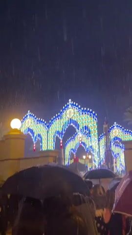 The video captures a nighttime scene amidst heavy rain. The camera is oriented upwards, focusing on illuminated decorative arches adorned with blue and white lights. These arches stand in front of two tall, yellow and red towers, one of which is topped with a statue. Below, a crowd of people is visible, holding up umbrellas to shield themselves from the downpour.  The combination of festive lighting and inclement weather creates a juxtaposition of celebration and challenge.