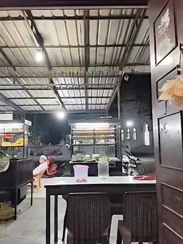 The video shows the interior of a restaurant at night.  The ceiling is a metal grid with lights.  There are tables and chairs.  A motorcycle is parked outside.  A food stall with rotating skewers of food is visible.  A person is seated in the background, wearing what appears to be a white head covering.  A pink container and a clear jar sit on a table.  The video appears to be taken from inside a building, looking out onto an open-air dining area. Pictures are visible on the wall in the foreground.