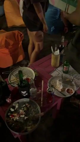 The video shows a night scene at a beachside establishment. In the foreground, a low table is cluttered with used plates, bottles of alcohol, and some food items. Further back, several people are seated on large, colorful beanbag chairs arranged on the sand, some under large umbrellas. The atmosphere appears relaxed and social, with people interacting and enjoying drinks. Trees with hanging lights provide ambiance.