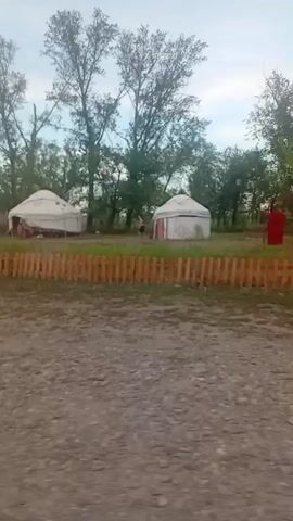 The video shows a campground with several yurts, wooden fences, trees, and a cloudy sky. In the foreground, there is a gravelly path. In the background, there are two white yurts and a red structure. There is a person walking near one of the yurts. The camera is moving, suggesting it is being filmed from a vehicle or a person walking.