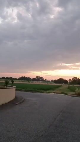 The video shows a rural landscape at dusk. A paved road curves to the right in the foreground. To the left, a low, curved wall with a few plants is visible. Beyond the road, there is a large field of green grass, bordered by trees and vegetation. The sky is filled with dramatic clouds, illuminated by the setting sun which casts a warm glow across the horizon. The light suggests the sun is low in the sky, likely setting. There are no people or animals visible in the video. The overall impression is one of peaceful evening scenery.