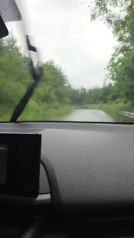 The video shows a view from inside a car driving on a wet road through a wooded area. The windshield wipers are intermittently wiping the rain from the glass. The road is narrow and appears to be paved, winding through dense green trees on either side. The sky is overcast and the overall lighting suggests a cloudy or rainy day. The dashboard of the car is visible in the foreground, with a dark electronic screen on the left side. The camera is stationary within the car, capturing the forward view as the vehicle moves.