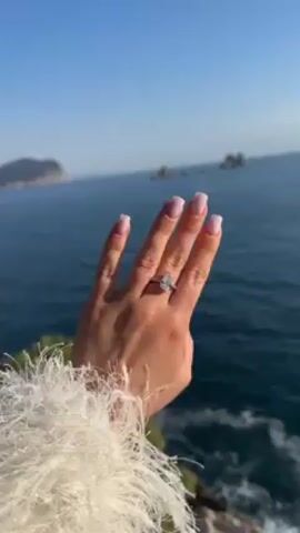 A hand with an engagement ring is held up in front of a scenic ocean view. The hand is tanned, with manicured nails painted light pink. A large diamond ring is on the ring finger. The person is wearing a fluffy, white garment. Behind the hand is a blue ocean, a rocky coastline, and a clear, blue sky.