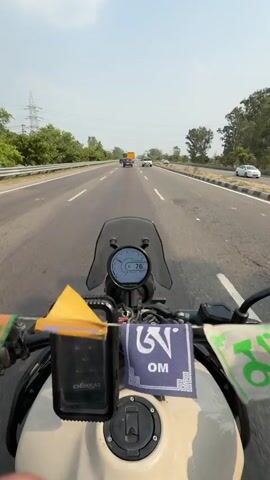 This video depicts a motorcycle ride on a highway. The camera is mounted on the handlebars, offering a first-person perspective of the journey. The rider, wearing a red and black helmet, black riding jacket, and gloves, is visible in the foreground. The motorcycle's dashboard displays a speed of 76.

The highway appears to be a multi-lane road with other vehicles, including cars, in the distance. The surrounding landscape is characterized by lush green trees and vegetation, suggesting a rural or semi-rural setting. The sky is bright and appears to be a clear day, indicative of daytime. The presence of power lines overhead also suggests infrastructure development alongside the natural scenery.

Decorative prayer flags, featuring Tibetan script, are attached to the motorcycle's handlebars. One flag has the mantra "Om Mani Padme Hum" in blue Tibetan script. The overall atmosphere conveys a sense of travel and exploration. The visible text on the rider's t-shirt is difficult to read clearly due to the resolution and angle, but it appears to be in English letters. The location context provided is Nilokheri, India.