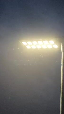 The video shows a brightly lit street light at night. The light fixture is rectangular and contains approximately twelve individual LED bulbs, all glowing with a warm white or yellowish light. Numerous small insects, likely moths, are seen fluttering rapidly around the light, attracted to its illumination. The street light is mounted on a tall, slender pole, which is visible on the right side of the frame. The background is a dark, indistinct sky, indicating nighttime conditions. The air around the light appears hazy or dusty due to the light illuminating airborne particles or the insects themselves. No people are visible, and no text is discernible. The setting is outdoors in what appears to be a street or public area.